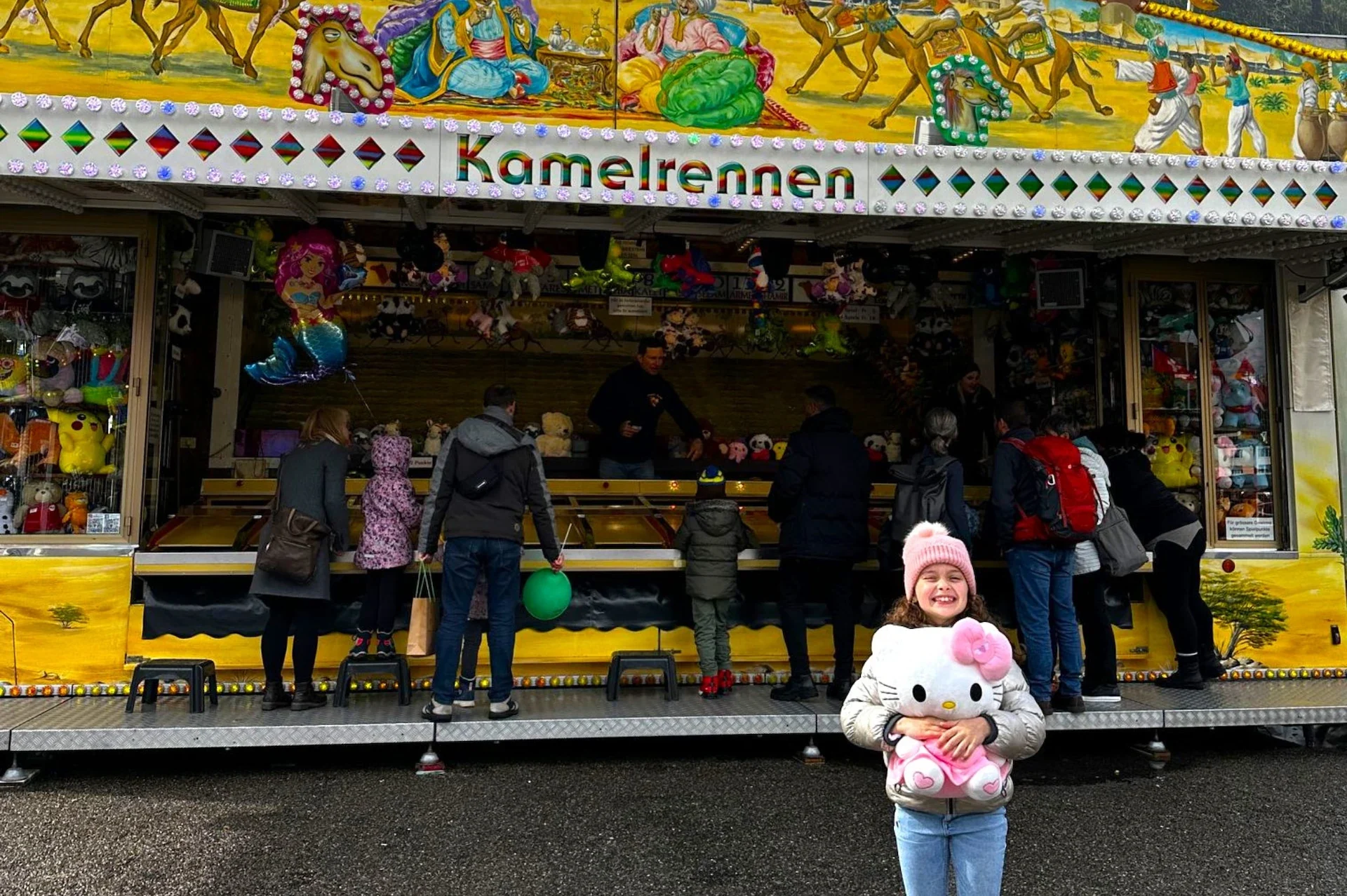 Kamelrennen Stand im Hintergrund, davor ein lachendes Mädchen mit Hello Kitty Plüschtier